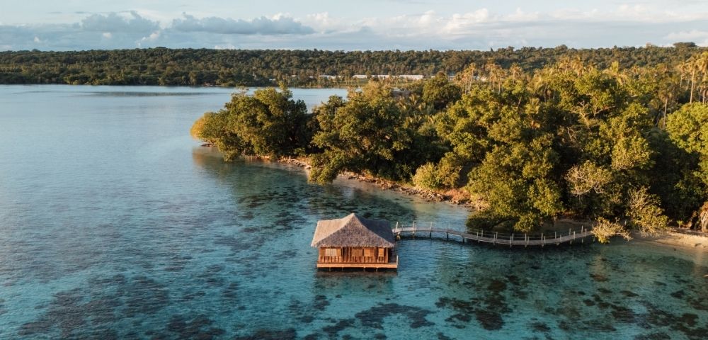 An overwater bungalow with a thatched roof stands in crystal-clear turquoise water, connected by a wooden boardwalk to a lush, green island coastline.