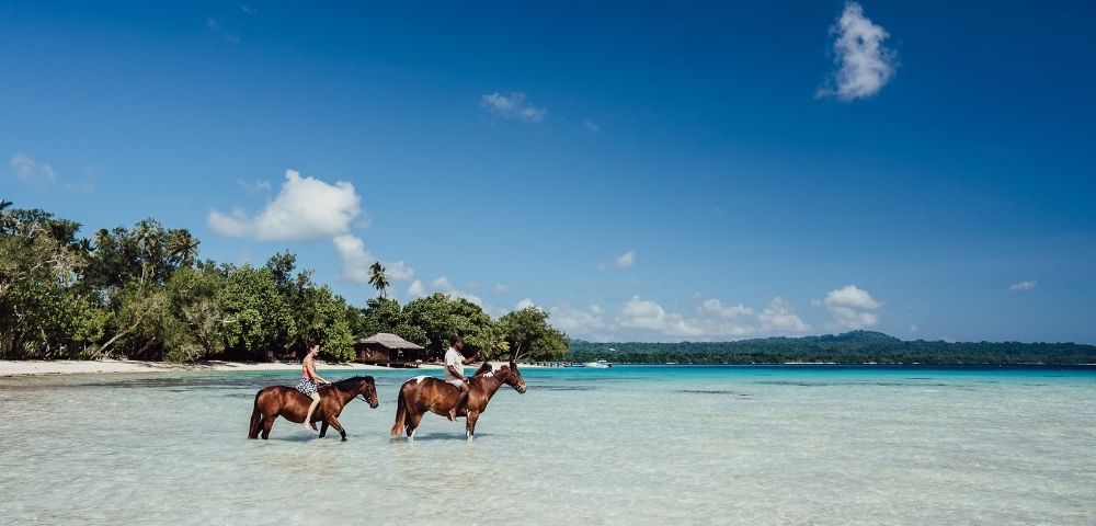 Two people ride horses through shallow, crystal-clear turquoise water on a tropical beach under a bright blue sky.