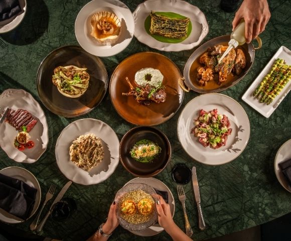 Overhead view of a dark green dining table set with colorful gourmet dishes as guests serve food, creating an elegant, convivial mood.