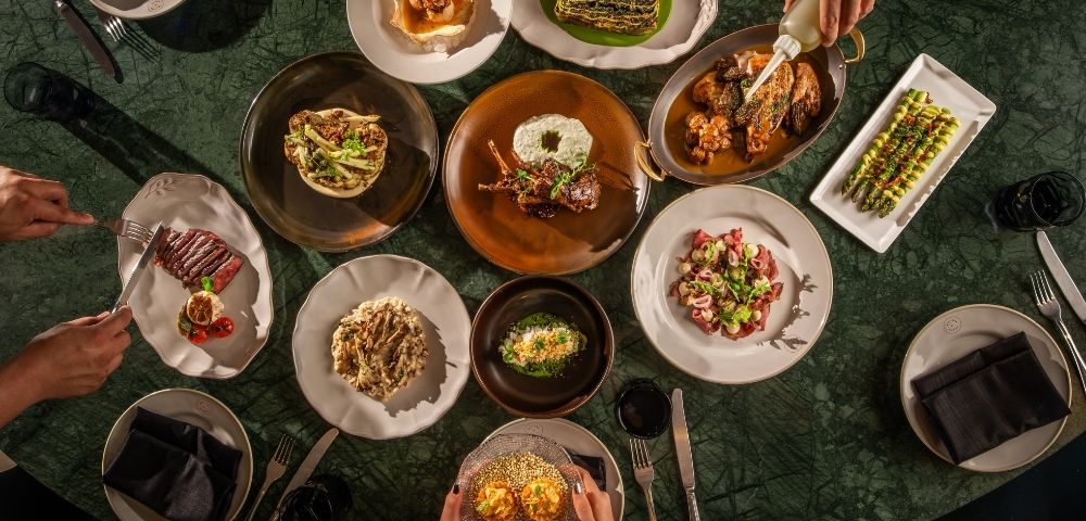Overhead view of a dark green dining table set with colorful gourmet dishes as guests serve food, creating an elegant, convivial mood.