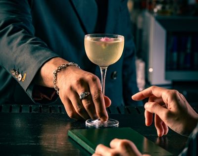 A bartender in a dark suit sets a stemmed cocktail on a black counter as a guest reaches toward it, creating an intimate, elegant bar mood.