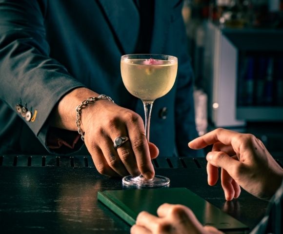 A bartender in a dark suit sets a stemmed cocktail on a black counter as a guest reaches toward it, creating an intimate, elegant bar mood.