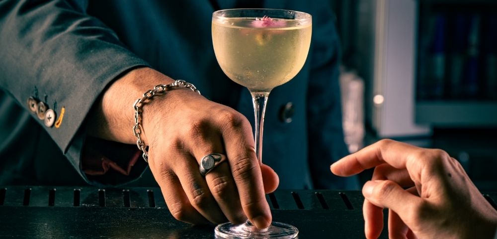A bartender in a dark suit sets a stemmed cocktail on a black counter as a guest reaches toward it, creating an intimate, elegant bar mood.