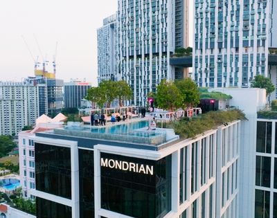 Rooftop pool and landscaped terrace atop a modern high-rise marked “MONDRIAN,” surrounded by dense city buildings and cranes under a bright sky.