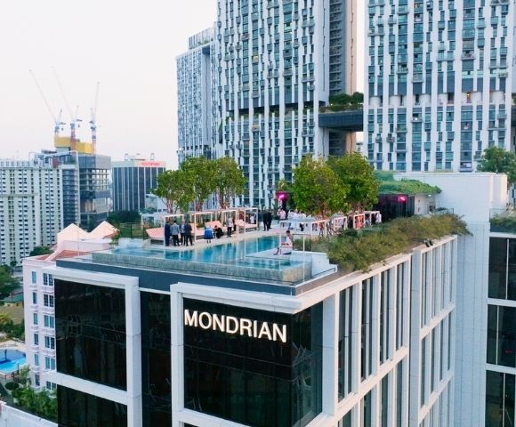Rooftop pool and landscaped terrace atop a modern high-rise marked “MONDRIAN,” surrounded by dense city buildings and cranes under a bright sky.
