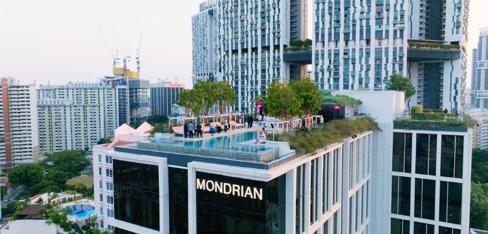Rooftop pool and landscaped terrace atop a modern high-rise marked “MONDRIAN,” surrounded by dense city buildings and cranes under a bright sky.