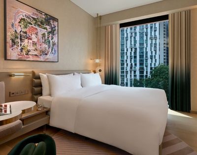 A serene, upscale hotel bedroom with a neatly made white bed, colorful framed artwork, a book on the bedside table, and a city view through tall windows.