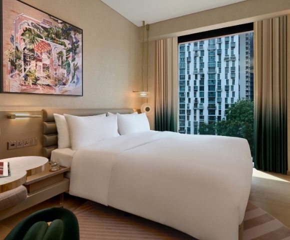 A serene, upscale hotel bedroom with a neatly made white bed, colorful framed artwork, a book on the bedside table, and a city view through tall windows.