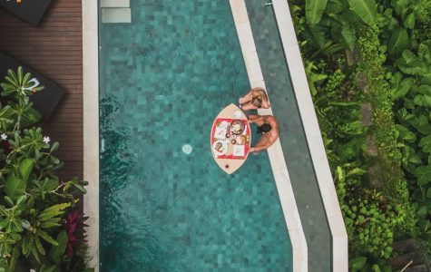 Aerial view of a pool surrounded by lush greenery and a deck. A couple relaxes on a floating tray with bright drinks, conveying a tranquil, tropical vibe.