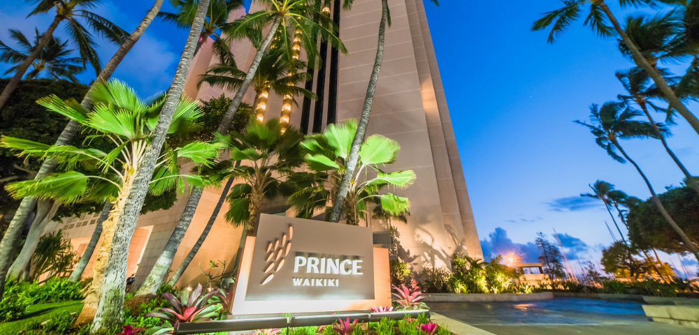 Exterior of the Prince Waikiki hotel at twilight, surrounded by palm trees and colorful foliage, with a clear blue sky in the background.
