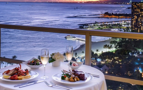 Romantic dinner setting on a balcony, overlooking a sunset-lit beach and city skyline, featuring seafood and gourmet dishes.