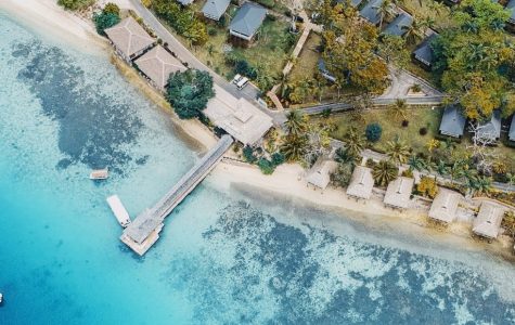 Aerial view of a peaceful tropical island, with turquoise water, anchored boats, a sandy beach, and small huts nestled among lush trees.