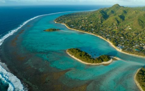 Aerial view of a tropical coastline with clear turquoise water, white sandy beaches, lush green islands, and a small village nestled amongst palm trees.