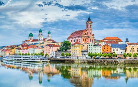 A scenic view of a European town by a river. Colorful buildings line the waterfront, with a prominent church and cloudy blue sky reflected in the water.