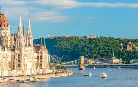 Panoramic view of Budapest with the Parliament building by the Danube River, lush green hills, and Chain Bridge on a sunny day. Boats dot the river.