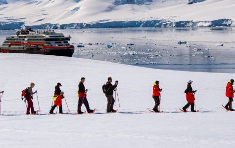 A group of people in red jackets hike in a line across a snow-covered landscape. A large ship floats on calm icy waters beneath snow-capped mountains.