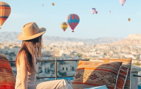 A woman in a hat sits on a rooftop terrace with colorful cushions, watching numerous hot air balloons float in a clear blue sky over a scenic valley.