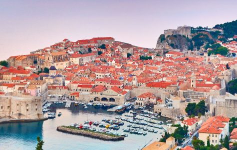 Panoramic view of Dubrovnik, Croatia. The historic city features orange-tiled roofs, stone walls, and a harbor with boats. Lush greenery surrounds the buildings.