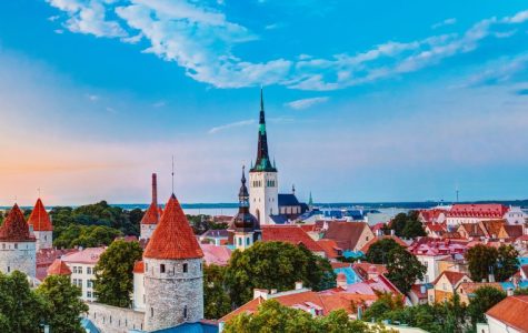 A picturesque view of Tallinn's skyline, featuring medieval towers with red roofs, a tall spire, lush greenery, and a blue sky at sunset. Tranquil and historic.