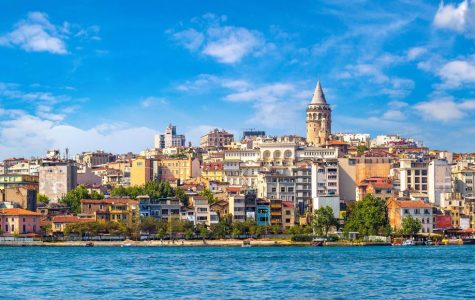 Scenic view of Istanbul's skyline with the historic Galata Tower rising above colorful buildings. A calm blue sea and a clear sky enhance the lively atmosphere.