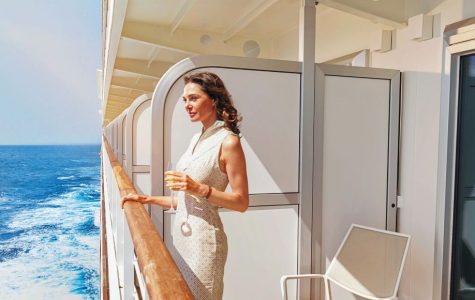 A woman in a white dress enjoys a drink on a cruise ship balcony, overlooking the sparkling ocean under a sunny sky. The scene is serene and luxurious.
