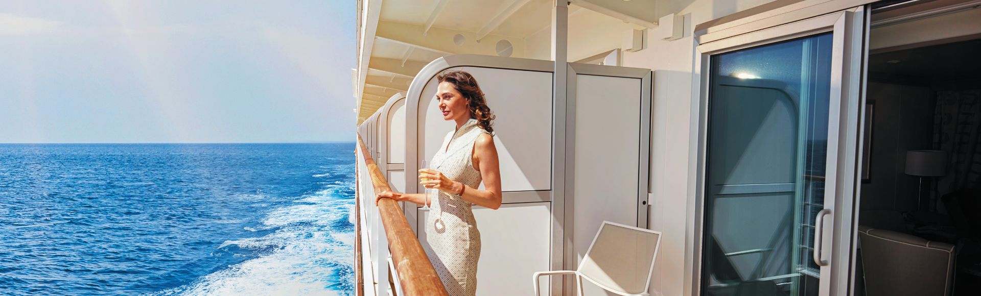 A woman in a white dress enjoys a drink on a cruise ship balcony, overlooking the sparkling ocean under a sunny sky. The scene is serene and luxurious.