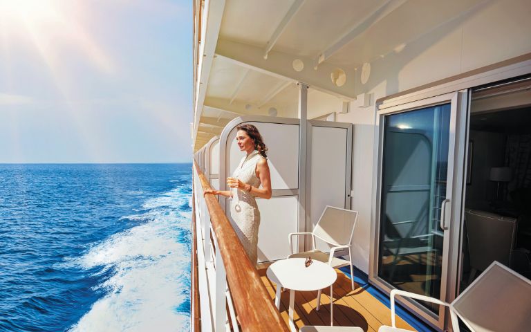 A woman in a white dress enjoys a drink on a cruise ship balcony, overlooking the sparkling ocean under a sunny sky. The scene is serene and luxurious.