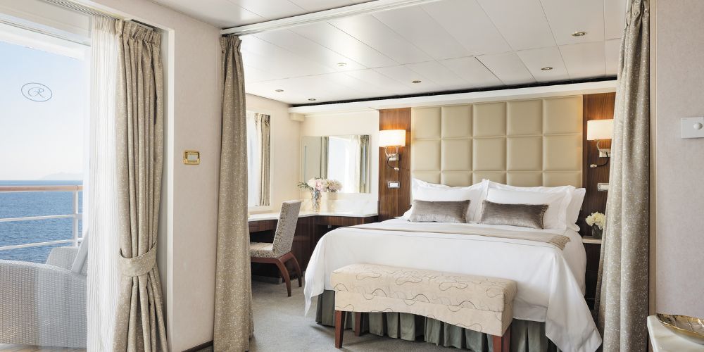 Luxurious cruise ship cabin with a large, elegant bed, plush pillows, and soft lighting. A window reveals a serene ocean view. Calm and inviting atmosphere.
