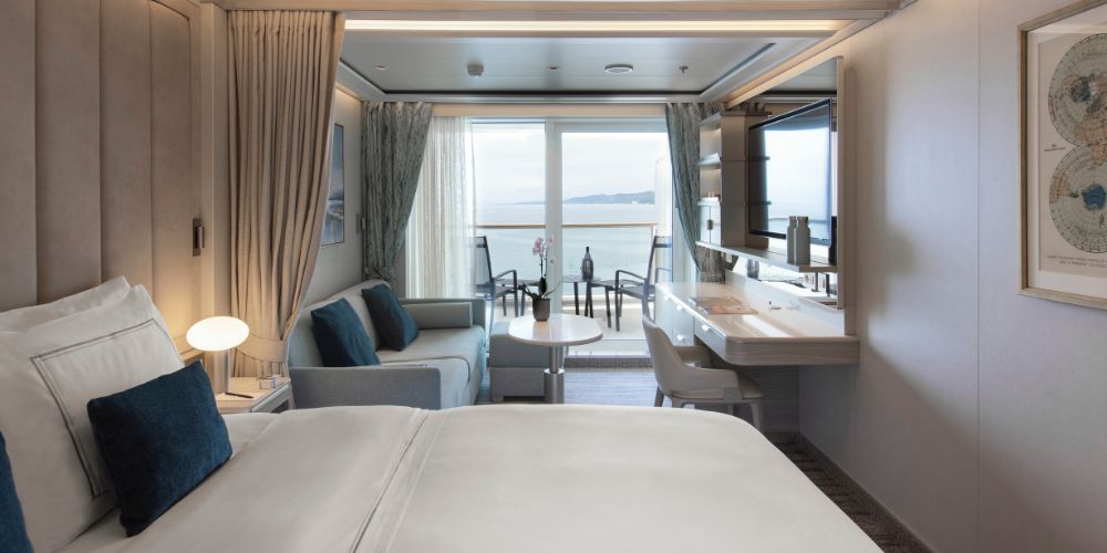 Modern hotel room with a large bed, sofa, and desk. Glass doors reveal a balcony overlooking a serene ocean view. Neutral tones create a calming ambiance.