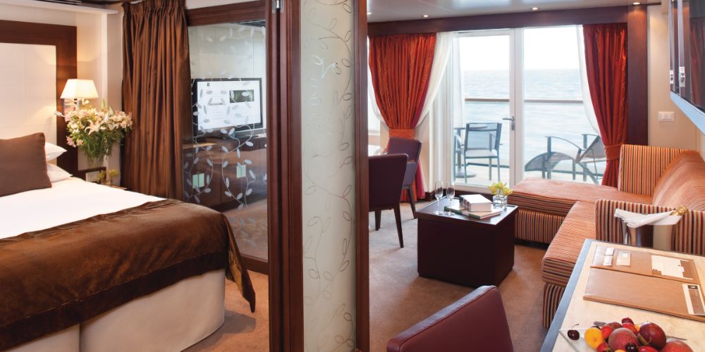 Luxurious cruise cabin with ocean view. Cozy bedroom has a plush bed, flowers, and soft lighting. Living area has a sofa, table, and balcony access for relaxation.