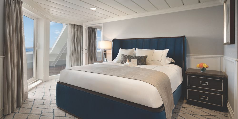 A cozy, elegant bedroom features a plush blue bed with white and beige bedding, flanked by a wooden nightstand and lamp. Large windows reveal a sunny balcony.