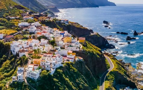 A picturesque coastal village with colorful houses atop a lush, green cliff. Mountains rise in the background, and the vast ocean stretches under a bright blue sky.