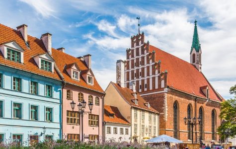 Historic European street scene with colorful gabled houses, a brick church with a green spire, and vintage lampposts under a vibrant blue sky.