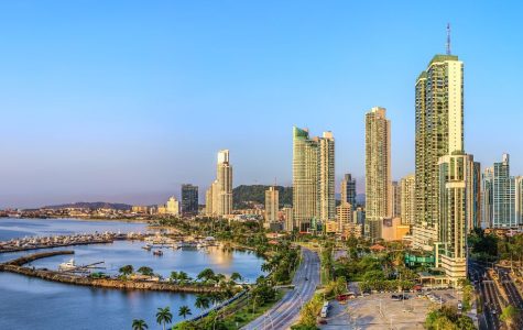 A panoramic view of a coastal city at sunset, showcasing tall skyscrapers, a curving shoreline, and palm-lined roads. The scene conveys a serene urban atmosphere.