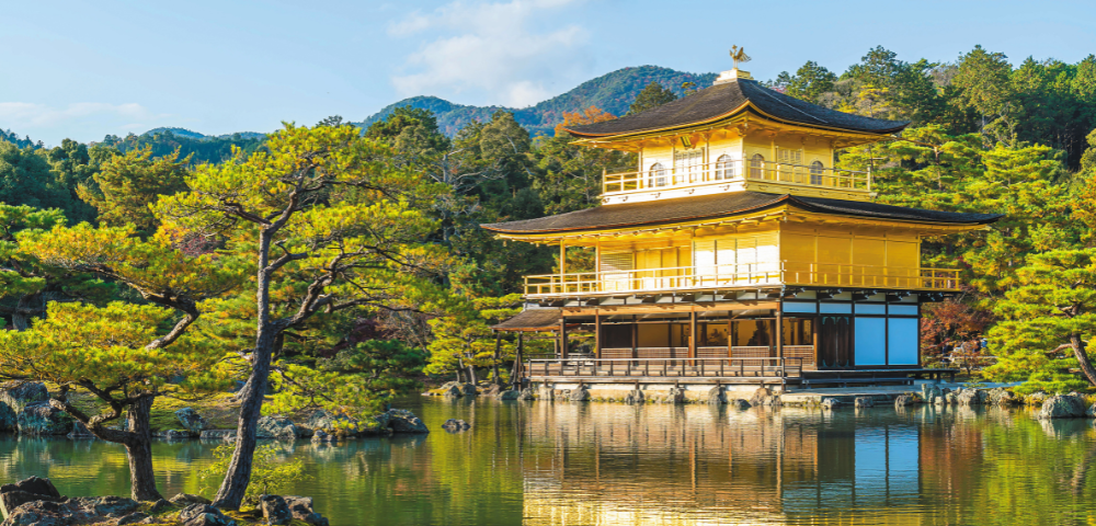 Golden Pavilion temple reflected in a calm pond, surrounded by pine trees and mountainous forest in the background.