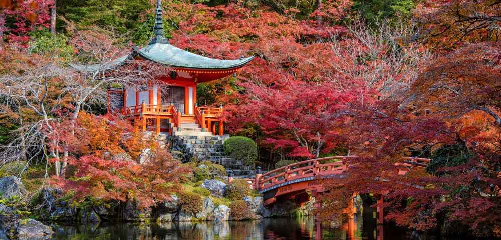 A serene Japanese garden in autumn features a traditional red pagoda and bridge surrounded by vibrant red and orange foliage, reflecting in a calm pond.