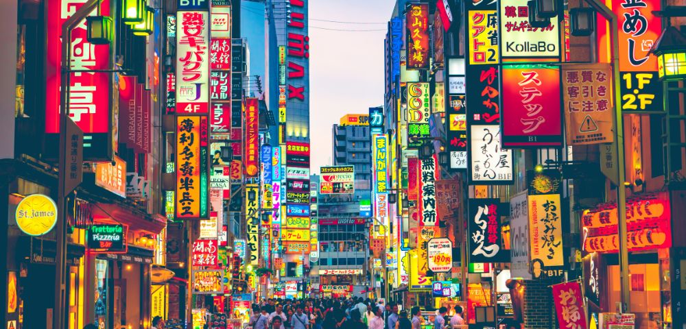 A vibrant city street in Tokyo, Japan, with colorful neon signs in Japanese and English. The bustling crowd creates a lively and dynamic atmosphere.