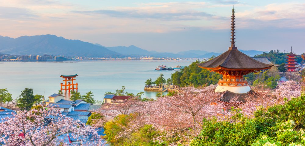 Scenic view of Itsukushima Shrine's torii gate on water, surrounded by cherry blossoms and traditional pagodas, with mountains in the background at sunset.