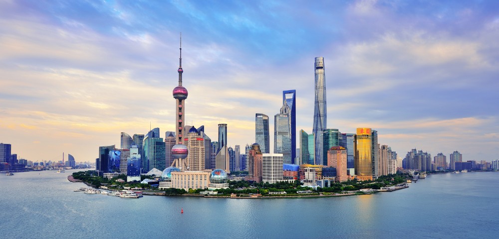 Skyline of Shanghai at sunset, featuring modern skyscrapers like the Oriental Pearl Tower by the waterfront. The sky is a mix of blue and orange hues.