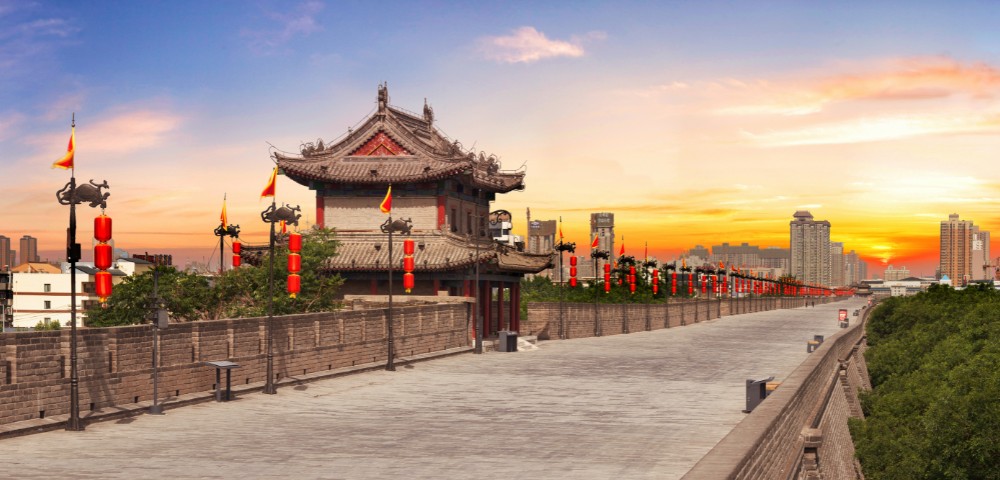 Ancient city wall with ornate tower, red lanterns, and flags under a vibrant sunset sky, creating a serene and historical ambiance.