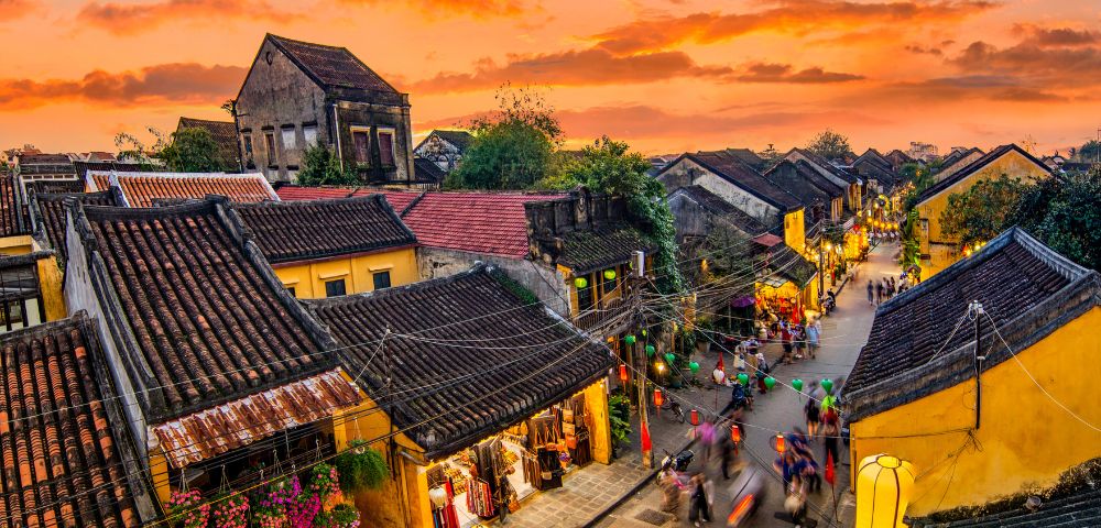 A vibrant street in an ancient town at sunset, with yellow lantern-lit buildings, bustling people, and a dramatic orange sky creating a lively atmosphere.