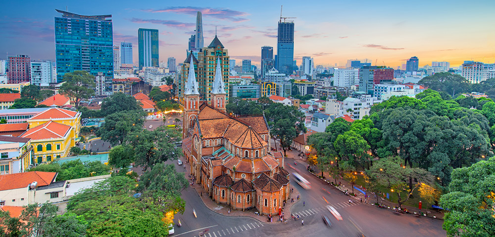 Aerial view of a vibrant cityscape at sunset, featuring a historic cathedral with twin spires amid lush trees and modern skyscrapers in the background.