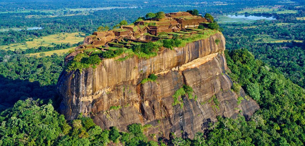 Aerial view of Sigiriya, a massive rock fortress in Sri Lanka. Ruins are atop the rock, surrounded by lush forests and distant landscapes. Majestic and historical.