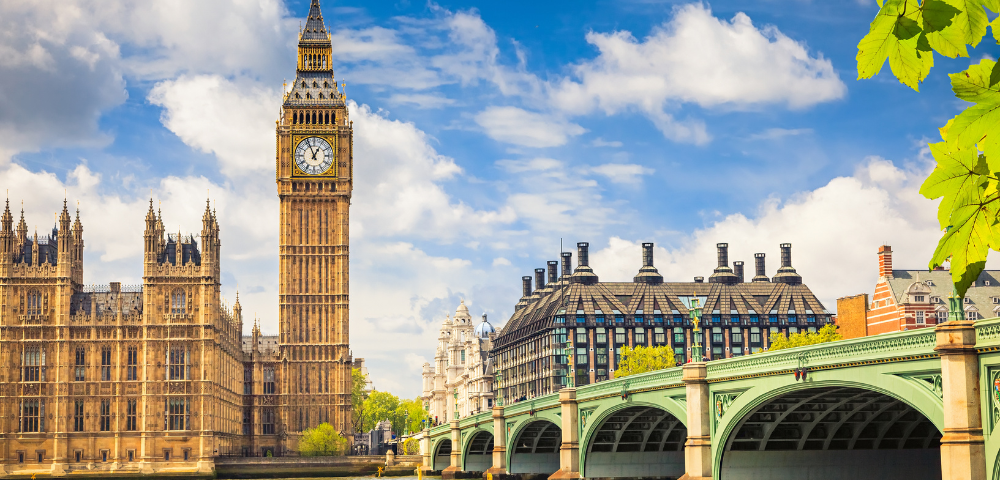Iconic clock tower beside the Houses of Parliament under a bright blue sky with white clouds. Nearby is a green bridge, lush greenery, and historic architecture.