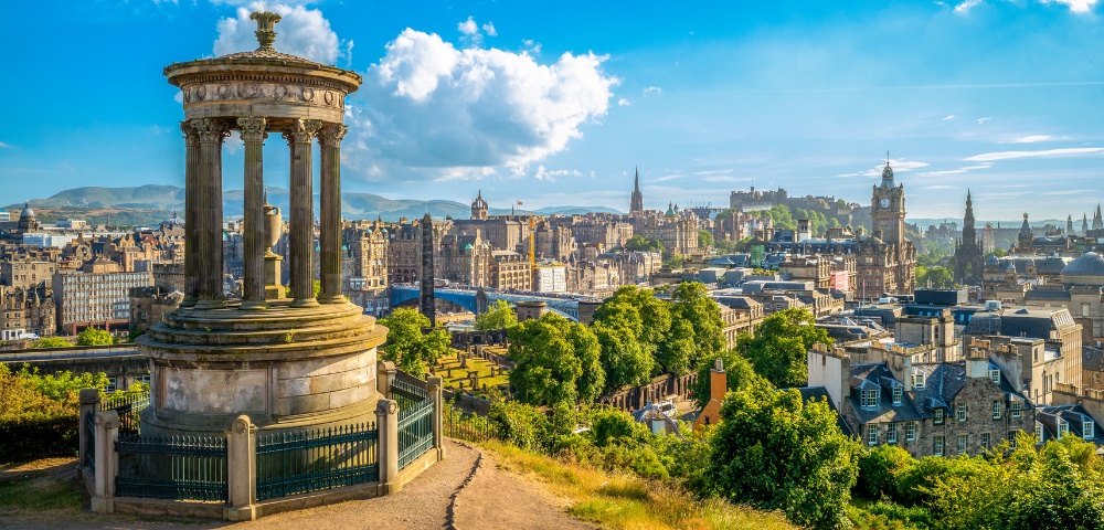 Panoramic view of Edinburgh cityscape with the Dugald Stewart Monument in the foreground under a bright blue sky.