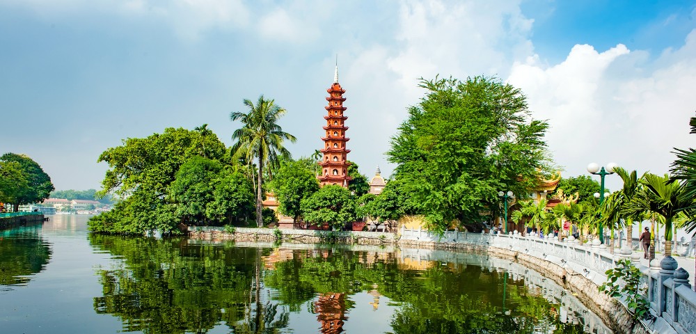 A red pagoda rises above lush green trees beside a calm lake, with a curved white stone railing pathway on the right under a partly cloudy sky.