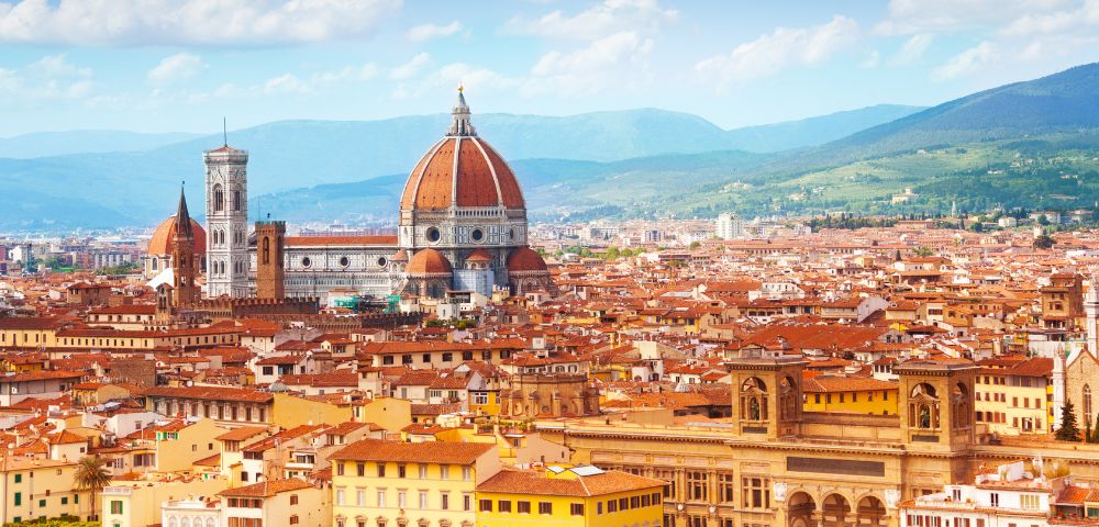 Panoramic view of Florence, Italy, featuring the red-domed Florence Cathedral and surrounding historic buildings under a blue sky.