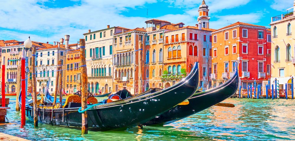 Two black gondolas docked on a canal in Venice with colorful historic buildings and blue sky in the background.