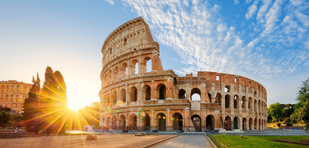 Sunset over the ancient Colosseum in Rome with sun rays shining through trees and a partly cloudy blue sky.