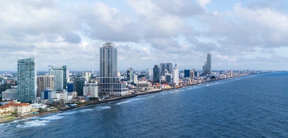 Aerial view of a coastal cityscape with skyscrapers lining the shore. The ocean is on the right, waves gently lapping. The sky is partly cloudy.